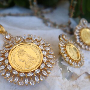 May include: A gold pendant necklace and earrings set with a round coin-shaped charm surrounded by clear cubic zirconia stones. The coin features a profile of a woman.