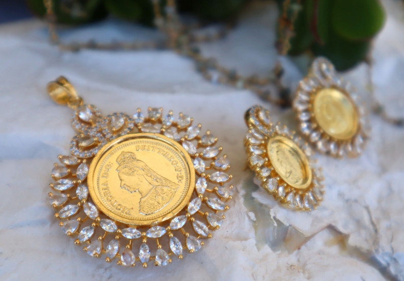 May include: A gold pendant necklace and earrings set with a round coin-shaped charm surrounded by clear cubic zirconia stones. The coin features a profile of a woman.