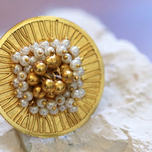 May include: A gold ring with a circular design and a cluster of white pearls and gold beads in the centre.
