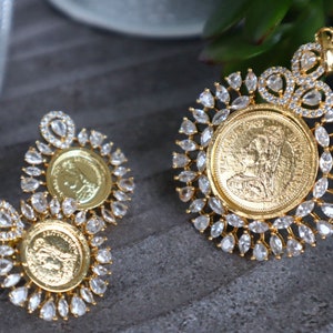 May include: A gold pendant and earrings set featuring a round coin design with a woman's profile. The coin is surrounded by a halo of clear, sparkling stones.