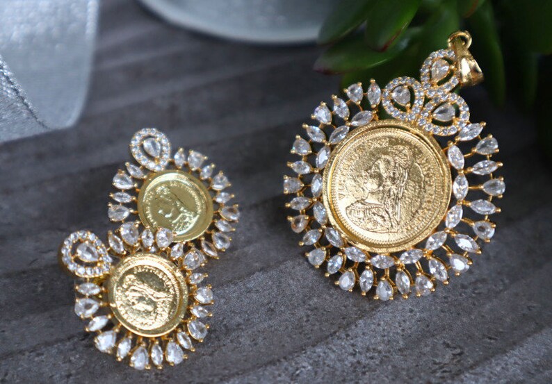 May include: A gold pendant and earrings set featuring a round coin design with a woman's profile. The coin is surrounded by a halo of clear, sparkling stones.