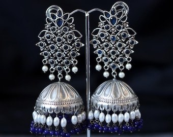 May include: A pair of silver-toned chandelier earrings with a detailed design. The earrings feature dark blue gemstones, white pearls, and a bell-shaped bottom. The earrings are displayed on a silver stand against a black background.