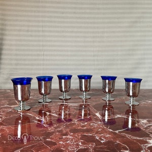 May include: Set of six silver-plated shot glasses with blue glass tops. The glasses are arranged in a row on a marble surface.
