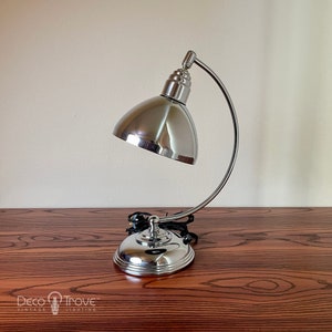 May include: A chrome desk lamp with a curved arm and a round, dome-shaped shade. The lamp is on a wooden table.