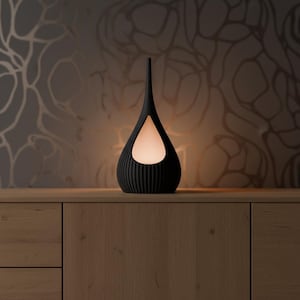 Könnte beinhalten: Eine schwarze, tropfenförmige Lampe mit vertikalem Rippendesign. Die Lampe hat einen langen, schlanken Hals und einen leuchtenden, weißen Innenraum, der durch eine tropfenförmige Öffnung sichtbar ist. Die Lampe steht auf einer Holzoberfläche.