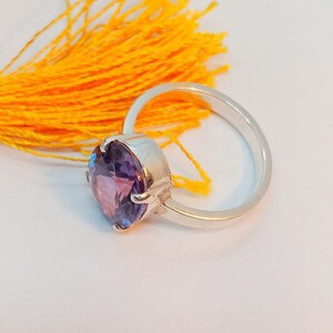 Puede incluir: Un anillo de plata con una gran piedra preciosa redonda de color p&uacute;rpura. El anillo est&aacute; colocado sobre un fondo amarillo.
