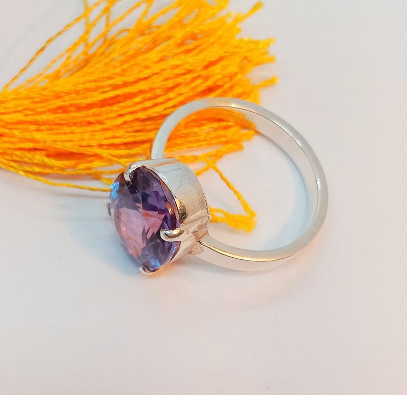 Puede incluir: Un anillo de plata con una gran piedra preciosa redonda de color p&uacute;rpura. El anillo est&aacute; colocado sobre un fondo amarillo.