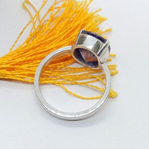 Puede incluir: Un anillo de plata con una piedra preciosa p&uacute;rpura. El anillo est&aacute; colocado sobre una borla amarilla.
