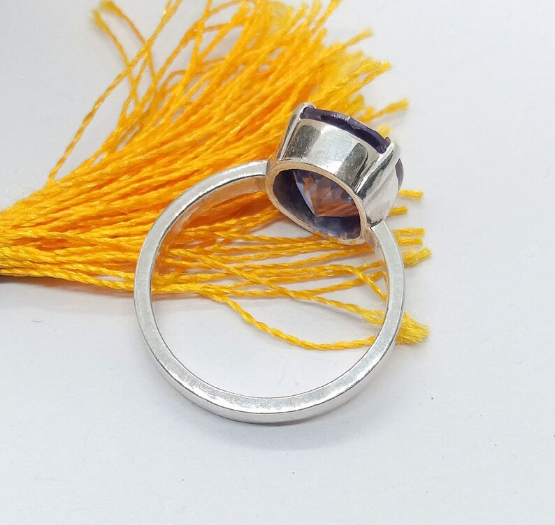Puede incluir: Un anillo de plata con una piedra preciosa p&uacute;rpura. El anillo est&aacute; colocado sobre una borla amarilla.