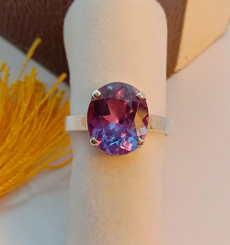Puede incluir: Un anillo de plata con una gran piedra preciosa de forma ovalada. La piedra preciosa es de color morado oscuro que se desvanece a un azul m&aacute;s claro en la parte inferior. El anillo est&aacute; engastado con cuatro peque&ntilde;as garras de plata.