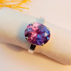 Puede incluir: Un anillo de plata con una gran piedra preciosa de forma ovalada. La piedra preciosa es de color morado oscuro con un tono rosado.