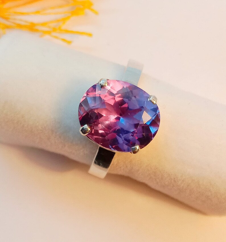 Puede incluir: Un anillo de plata con una gran piedra preciosa de forma ovalada. La piedra preciosa es de color morado oscuro con un tono rosado.