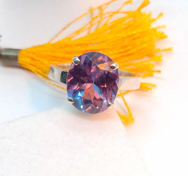 Puede incluir: Un anillo de plata con una gran piedra preciosa ovalada de color p&uacute;rpura y azul. La piedra preciosa tiene un corte facetado y est&aacute; engastada en una banda simple.