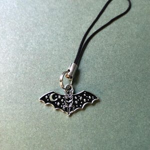 May include: A silver-coloured bat charm with a black enamel finish, featuring a crescent moon and star details. The charm is attached to a black cord with a silver-coloured metal clasp. The bat's wings are spread, and the design has a gothic aesthetic.