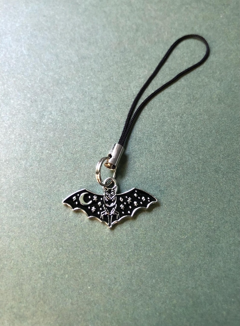 May include: A silver-coloured bat charm with a black enamel finish, featuring a crescent moon and star details. The charm is attached to a black cord with a silver-coloured metal clasp. The bat's wings are spread, and the design has a gothic aesthetic.