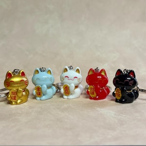 May include: Five Maneki-neko cat keychains in gold, light blue, white, red, and black. Each cat figure has a smiling face, pointed ears, and holds a small gold-colored plaque with Japanese characters. Each keychain includes a silver metal ring.