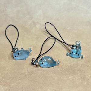 May include: Three translucent light blue seal charms made of glass or resin, each featuring black painted eyes, whiskers, and a nose. Every charm is attached to a thin black cord loop with a small silver-toned metal jump ring connector.