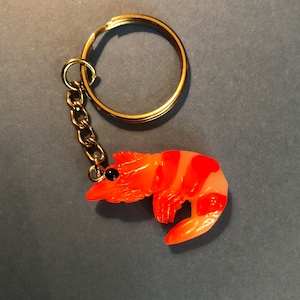 May include: A gold key chain with a bright orange and white shrimp charm. The shrimp charm has black eyes and is detailed with white stripes.