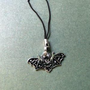 May include: A silver bat pendant with black enamel detailing, suspended from a black cord. The bat features detailed wing patterns and star accents. This pendant is likely a charm or accessory.