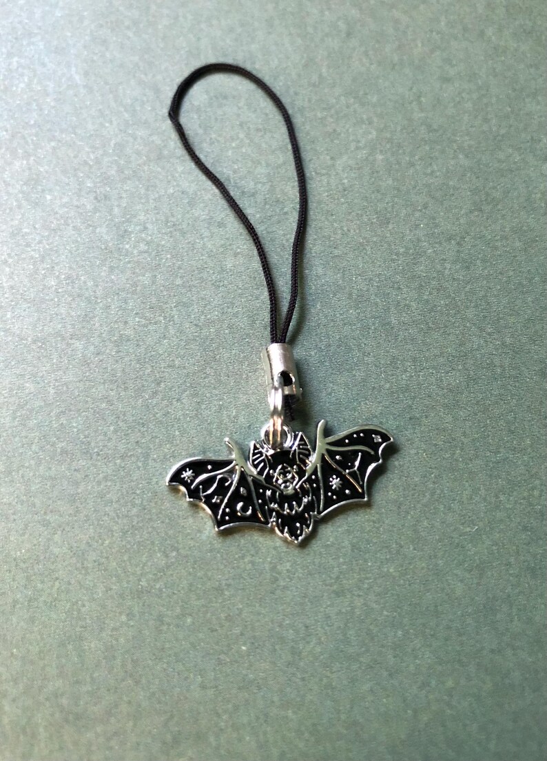 May include: A silver bat pendant with black enamel detailing, suspended from a black cord. The bat features detailed wing patterns and star accents. This pendant is likely a charm or accessory.