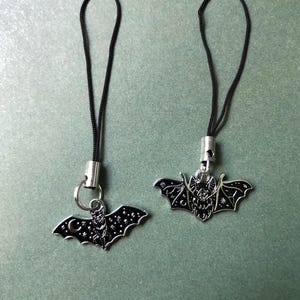 May include: Two silver-toned bat charms with black enamel detailing. Each charm has a black cord loop. The bats have intricate designs, including stars and a crescent moon. The charms are likely for jewelry or accessories.