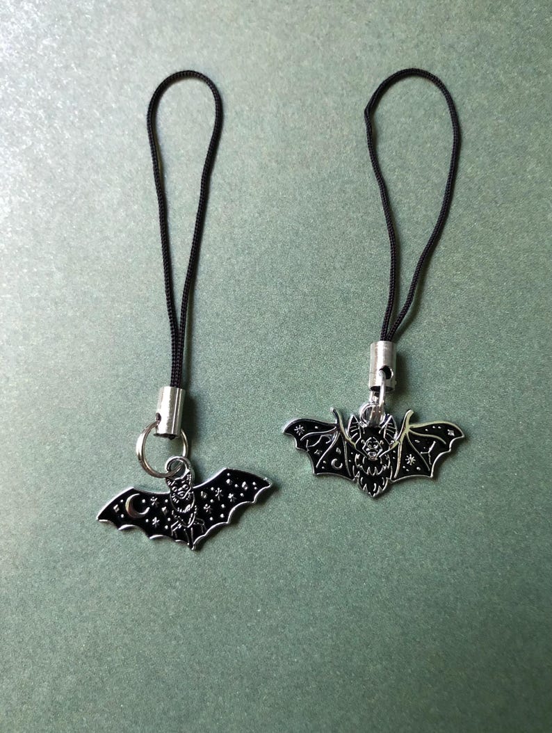 May include: Two silver bat charms with black enamel detailing. Each charm has a black cord loop. The bats feature intricate designs, including stars and a crescent moon. These charms are suitable for jewellery or accessories.