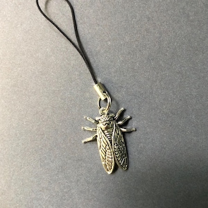 May include: A silver-colored cicada charm with detailed wings and body, attached to a black cord. The charm is suspended from a silver-toned metal loop and is set against a gray background. The cicada charm is a unique accessory.