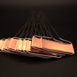 May include: A collection of copper-colored metal plant labels with a rectangular plate and a wire handle. The labels are arranged in a fan shape against a black background. The labels are for garden use.