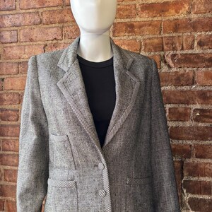 May include: A gray and black speckled blazer featuring a notched lapel with a small black and white checkered trim. The blazer includes three front buttons, two flap pockets, and a matching checkered border along the hem and pocket edges.