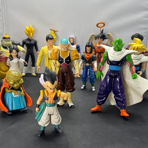 May include: A collection of twelve plastic action figures featuring various characters with distinct outfits, including a green-skinned figure in a white cape, a small figure in a blue skirt and orange cape, and several others in colorful attire.