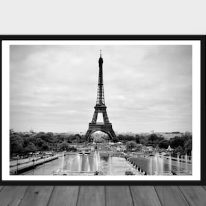 May include: A black and white photograph of the Eiffel Tower in Paris, France. The tower is in the center of the image, with a fountain in the foreground and trees and buildings in the background.