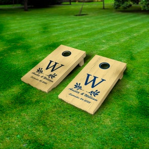 Op de afbeelding: Twee houten cornhole-borden met marineblauwe letters en ontwerp. Elk bord heeft een cirkelvormig gat en is gepersonaliseerd met de namen "Amelia & William" en de datum "September 24, 2022". De borden liggen op een groen gazon.