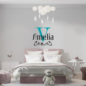 May include: A teal blue letter V with the name Amelia in black text below it. The name is decorated with a black floral design. The wall decal is on a white wall above a pink bed with white bedding and a white teddy bear.