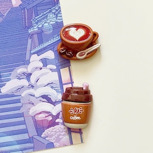 Op de afbeelding: Een bruine keramische koffiekop met een hartvormig ontwerp in het schuim, een kleine lepel en koffiebonen. Een bruine to-go koffiebeker met het woord "coffee" is ook zichtbaar. De achtergrond heeft een paars-blauwe illustratie.