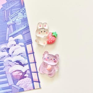 以下が含まれることがあります： 2つの樹脂チャーム：ピンクのイチゴを持った透明なウサギとピンクの猫。ウサギは黒い目を持ち、イチゴは緑の茎を持っています。猫はピンクのハートと黒いディテールを持っています。紫と白のイラストが背景にあります。