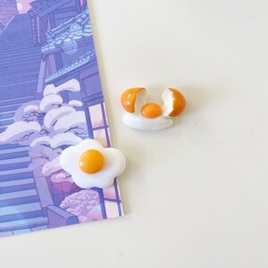 以下が含まれることがあります： セラミック製の目玉焼きのイヤリング2つ。片方のイヤリングは丸ごとの卵、もう片方は割れた卵で、黄身が見えています。