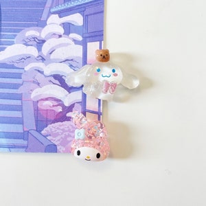 以下が含まれることがあります： 漫画のキャラクターの小さな透明なプラスチック製のフィギュア2つ。最初のフィギュアは白で、上に茶色のクマの頭とピンクのリボンが付いています。2番目のフィギュアはピンクで、頭に青い花とピンクのリボンが付いています。