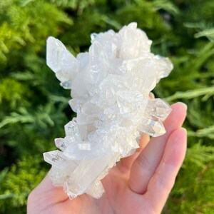 May include: A cluster of clear quartz crystals, held in a hand, with a natural, rough texture. The crystals are translucent and reflect light, showcasing their unique formations. The background is a blurred green, suggesting an outdoor setting.