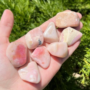 May include: Seven pink and white tumbled opal stones, some with a more pronounced pink hue, in a hand.