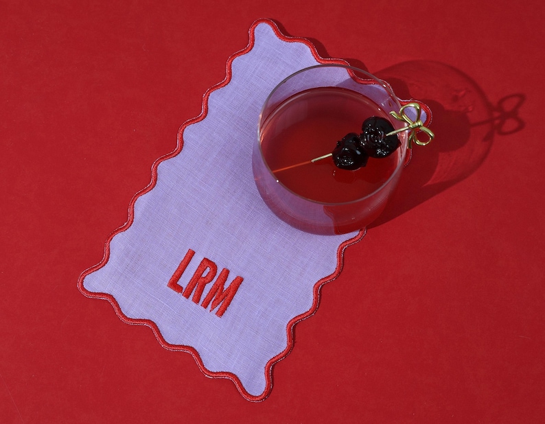 May include: A cocktail with dark cherries on a gold pick, resting on a lavender and white scalloped edge linen coaster with the embroidered letters "LRM". The coaster sits on a red surface.