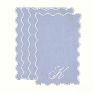 May include: A stack of light blue linen napkins with a white scalloped edge. Each napkin has a white embroidered letter "K" in the corner. The napkins are rectangular in shape.