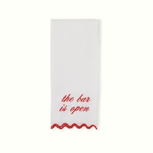 May include: A rectangular white hand towel featuring the red embroidered text "the bar is open" in a cursive script. The bottom edge of the towel is finished with a decorative red scalloped trim, providing a distinct contrast to the white fabric.