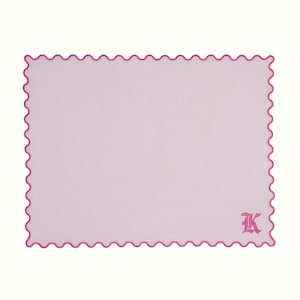 Op de afbeelding: Een lichtroze rechthoekige babydeken met een geschulpte rand in een donkerdere roze kleur. De deken heeft een geborduurde letter "K" in de rechter benedenhoek. De achtergrond is wit.