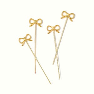 May include: Four gold-colored bow-shaped cocktail picks. The bows are at the top of the slender picks. The picks are arranged on a white background.