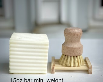 Puede incluir: Una barra de jabón blanca con textura acanalada, junto a un cepillo de fregar de madera con cerdas naturales. La barra de jabón está etiquetada con "15oz bar min. weight".