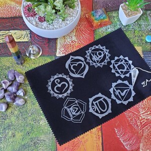 May include: A black felt mat with seven white chakra symbols printed on it. A clear quartz pendulum hangs above the mat. There are amethyst crystals, a rainbow-colored crystal wand, and a small bell on the table.