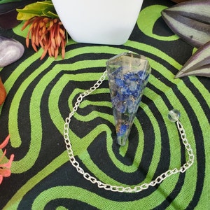 May include: A clear crystal pendulum with a silver chain and a blue and gold crystal point. The pendulum is on a green and black patterned surface.