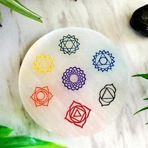 May include: White selenite charging plate with seven chakra symbols in different colors. The symbols are arranged in a circle on the plate. The plate is round and has a smooth surface.