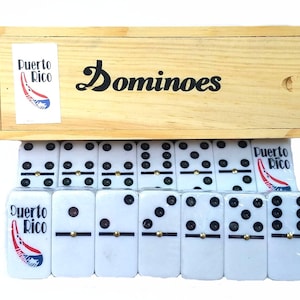 May include: A wooden box set of dominoes with the word "Dominoes" in black script. The box has a sticker that says "Puerto Rico" with a graphic of the island. The dominoes are white with black dots and gold accents.