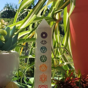 May include: White crystal point with seven chakra symbols in rainbow colors. The symbols are printed on the front of the crystal point. The crystal point is sitting on a green and black patterned surface.